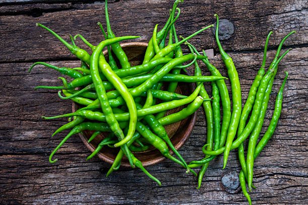 Fresh organic green chili peppers placed in a wooden bowl with some scattered around on rustic wooden surface.